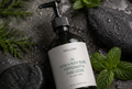 Peppermint and Dark Cedar hand and body wash surrounded by fresh mint leaves and wet spa stones, natural moisturizing body cleanser.